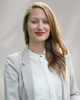 Eine lächelnde Frau mit langen, lockigen Haaren trägt einen hellen Blazer und ein Hemd. Der Hintergrund ist neutral und unauffällig.