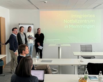 Fünf Personen stehen in einem Konferenzraum vor einer Präsentation mit dem Titel „Integriertes Notfallzentrum in Memmingen“. Tische und Stühle sind im Hintergrund sichtbar.