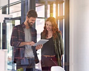 Psychologie in Organisationen | SRH Fernhochschule Zwei Personen stehen in einem modernen Büro und betrachten gemeinsam ein Tablet. Der Mann trägt ein kariertes Hemd, die Frau eine Jacke. Auf dem Tisch sind Unterlagen und Tassen sichtbar.
