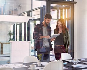 Psychologie in Organisationen | SRH Fernhochschule Zwei Personen stehen in einem modernen Büro und betrachten gemeinsam ein Tablet. Der Mann trägt ein kariertes Hemd, die Frau eine Jacke. Auf dem Tisch sind Unterlagen und Tassen sichtbar.