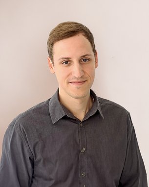 Ein junger Mann mit kurzen, braunen Haaren trägt ein graues Hemd und steht vor einer hellen Wand. Er lächelt leicht und schaut direkt in die Kamera.