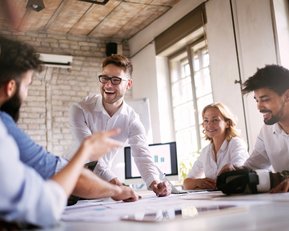 Eine Gruppe von fünf Personen in einem Besprechungsraum diskutiert und arbeitet an Dokumenten. Ein Mann präsentiert aktiv, während die anderen aufmerksam zuhören und Notizen machen.