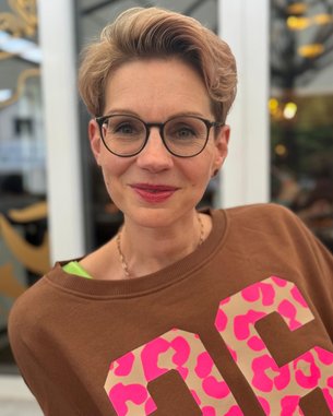 Frau mit kurzen, blonden Haaren und Brille trägt einen braunen Pullover mit großen, pinken Zahlen. Sie lächelt und steht vor einem hellen Hintergrund mit unscharfen Details.