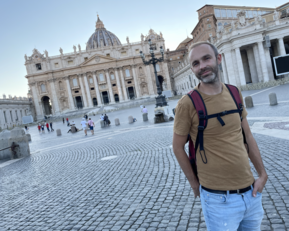 Ein Mann mit Bart steht auf dem Petersplatz in Rom, vor der beeindruckenden Fassade der Peterskirche. Er trägt ein braunes T-Shirt und eine Umhängetasche, während Menschen im Hintergrund spazieren.