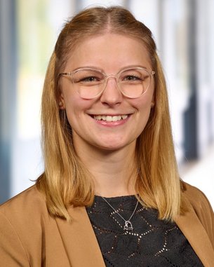 Eine lächelnde Frau mit blonden Haaren und Brille trägt eine beige Jacke und ein schwarzes Oberteil. Der Hintergrund zeigt unscharfe Fenster und Wände.