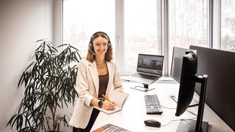Eine Frau mit Headset sitzt an einem Schreibtisch und arbeitet am Computer, während sie in einem modernen Büro umgeben von Pflanzen ist.