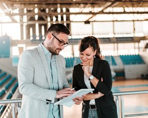 BWL und Sportmanagement Bachelor Ein Mann und eine Frau stehen in einem Sportstadion und betrachten gemeinsam ein Tablet. Beide scheinen in ein Gespräch vertieft zu sein, während im Hintergrund leere Sitzplätze sichtbar sind.