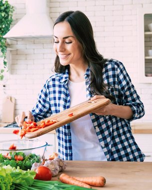 Eine Frau schneidet frisches Gemüse auf einem Holzbrett in einer modernen Küche. Sie lächelt, während sie die geschnittenen Zutaten in eine Schüssel gibt.
