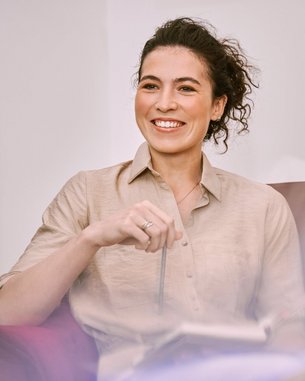 Eine Frau mit lockigem Haar sitzt entspannt in einem roten Sessel, hält einen Stift in der Hand und lächelt. Sie trägt ein beiges Hemd und hat ein Notizbuch auf ihrem Schoß.