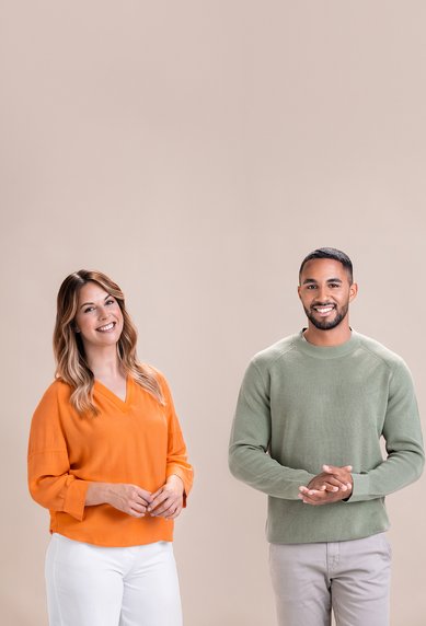 Zwei Personen stehen nebeneinander vor einem einfarbigen Hintergrund. Die Frau trägt einen orangefarbenen Pullover und die Hose ist weiß, während der Mann einen grünen Pullover und graue Hosen trägt.