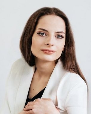 Eine Frau mit langen, braunen Haaren und einem neutralen Ausdruck trägt einen weißen Blazer und posiert vor einem hellen Hintergrund.