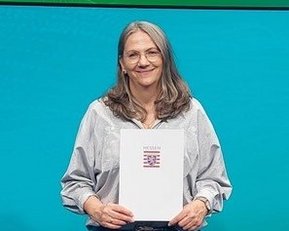 Eine Frau hält eine Urkunde in der Hand und steht vor einem Bildschirm mit dem Text "formnext". Sie trägt ein graues Oberteil und Jeans.