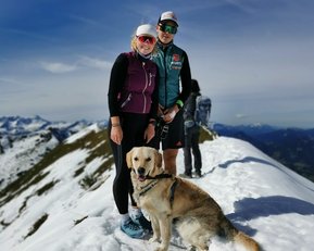 Zwei Personen stehen auf einem schneebedeckten Gipfel, umgeben von Bergen. Eine Person trägt eine Sonnenbrille und eine Weste, die andere einen Sportanzug. Ein Labrador sitzt vor ihnen.
