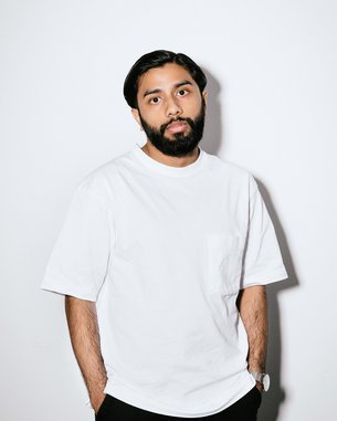 Ein Mann mit Bart trägt ein weißes T-Shirt und schwarze Hose. Er steht vor einem neutralen Hintergrund, die Hände lässig in den Hosentaschen.