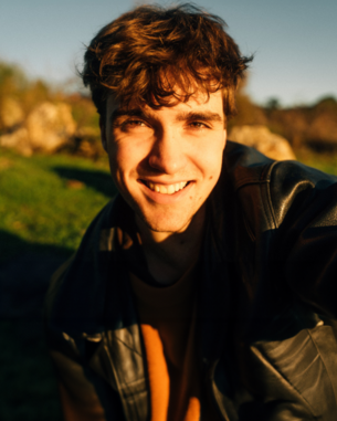 Ein junger Mann mit lockigem Haar lächelt in die Kamera, während er einen schwarzen Lederjacke und ein braunes Oberteil trägt.