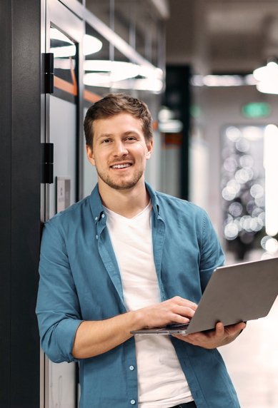 Ein Mann steht in einem modernen Büroflur und hält einen Laptop in der Hand. Er trägt ein hellblaues Hemd und lächelt.