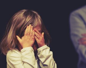 Ein kleines Mädchen mit schulterlangen Haaren hält sich die Hände vor das Gesicht, während zwei Personen im Hintergrund stehen.