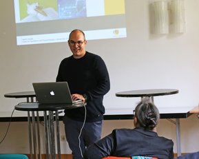 Ein Mann steht an einem Tisch mit einem Laptop und präsentiert vor einem Publikum in einem hellen Raum.
