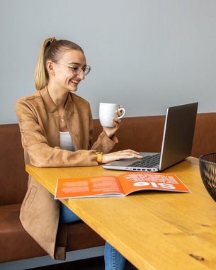 Eine Frau sitzt an einem Tisch, arbeitet am Laptop und hält eine Tasse in der Hand. Auf dem Tisch liegt eine Zeitschrift und eine Schüssel mit Obst.