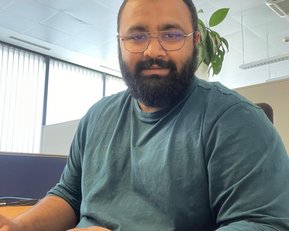 Ein Mann mit Bart und Brille sitzt an einem Schreibtisch und tippt auf einer Tastatur. Er trägt ein grünes Oberteil und hat eine Pflanze im Hintergrund. Tageslicht strömt durch große Fenster.