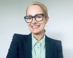 Eine Frau mit blonden Haaren trägt eine schwarze Blazerjacke und ein gestreiftes Hemd. Sie lächelt und hat die Arme verschränkt.