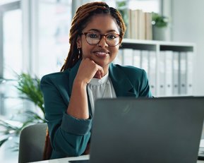 Eine Frau mit Brille und Dreadlocks sitzt an einem Schreibtisch und schaut lächelnd auf einen Laptop. Pflanzen sind im Hintergrund sichtbar.