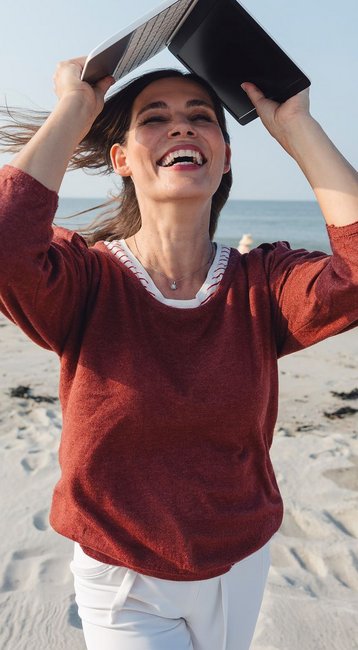 Eine Frau läuft am Strand und hält ein Buch über ihrem Kopf, während sie lächelt und die Meereswellen im Hintergrund sichtbar sind.