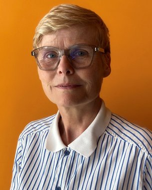Eine Frau mit kurzen, blonden Haaren und einer gestreiften Bluse steht vor einem orangefarbenen Hintergrund. Sie trägt eine modische Brille.