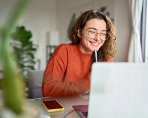 Eine lächelnde Frau mit Brille sitzt an einem Tisch, arbeitet am Laptop und hält einen Stift in der Hand. Ein Smartphone liegt daneben.