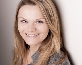 Eine lächelnde Frau mit langen, blonden Haaren steht vor einer hellen Wand. Sie trägt eine graue Jacke und einen orangefarbenen Oberteil.