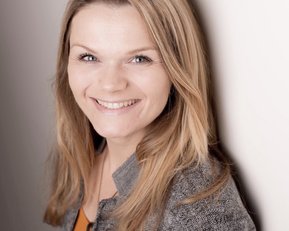 Eine lächelnde Frau mit langen, blonden Haaren steht vor einer hellen Wand. Sie trägt eine graue Jacke und einen orangefarbenen Oberteil.