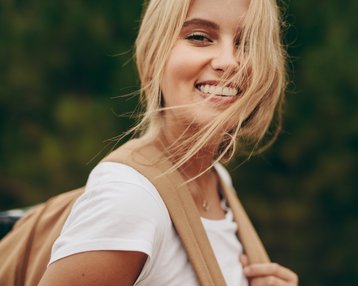 Junge Frau mit blonden Haaren und lächelndem Gesicht, trägt ein weißes T-Shirt und eine beige Tasche. Der Hintergrund zeigt verschwommenes Grün.