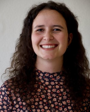 Eine lächelnde Frau mit lockigem, braunem Haar trägt ein blumiges Oberteil und eine Halskette mit einem Symbol. Der Hintergrund ist neutral und hell.