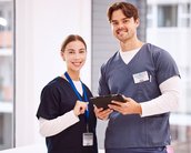 Zwei medizinische Fachkräfte stehen in einem hellen Raum, lächeln und halten ein Tablet. Beide tragen Arbeitskleidung und Namensschilder.