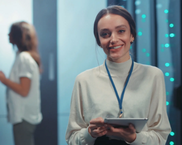 Eine lächelnde Frau mit einem Tablet in der Hand, trägt eine Bluse und eine Halskette, steht im Vordergrund, während eine andere Person im Hintergrund steht.
