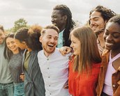 Eine Gruppe von zehn Personen steht eng beieinander und lächelt, während sie sich umarmen. Sie zeigen eine Vielfalt an ethnischen Hintergründen und tragen legere Kleidung. Der Hintergrund zeigt eine grüne Wiese und einen bewölkten Himmel.