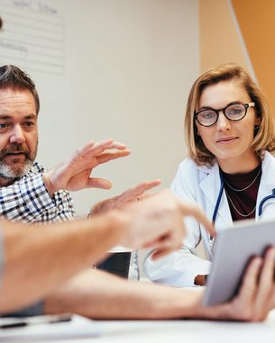 Vier Personen diskutieren an einem Tisch, während eine Frau in einem weißen Kittel ein Tablet betrachtet. Sie scheinen Informationen auszutauschen und zu analysieren.