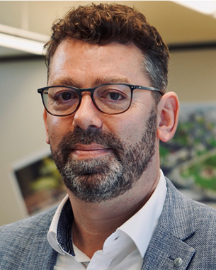 Ein Mann mit Brille und Bart trägt einen grauen Anzug und ein weißes Hemd. Er steht in einem modernen Raum mit einem Plan oder einer Karte im Hintergrund.