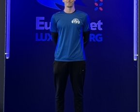 Ein junger Mann steht in einem blauen T-Shirt und schwarzen Hosen vor einem Hintergrund mit dem Logo von Eurobet Luxembourg.