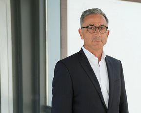 Ein Mann in einem Anzug steht vor einer modernen Glaswand. Er trägt eine Brille und hat graue Haare.