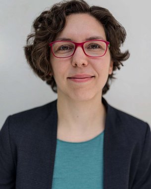 Eine Person mit lockigem, braunem Haar und roten Brillen sitzt vor einem hellen Hintergrund. Sie trägt ein dunkelblaues Blazer und ein türkisfarbenes Oberteil.