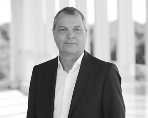 Ein Mann in einem schwarzen Anzug steht vor einer hellen, modernen Architektur. Er hat kurzes, graues Haar und lächelt leicht.