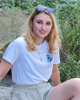 Eine junge Frau mit blonden Haaren sitzt auf einem großen Stein. Sie trägt ein weißes T-Shirt, beige Shorts und hat eine rote Bandage am Arm. Im Hintergrund sind grüne Pflanzen und Felsen sichtbar.