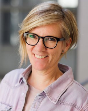 Eine lächelnde Frau mit blonden, schulterlangen Haaren und Brille trägt ein helles Hemd. Der Hintergrund ist unscharf und zeigt einen modernen Innenraum.