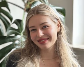 Eine junge Frau mit langen, blonden Haaren lächelt in die Kamera, während sie vor einer Fensterbank mit einer Pflanze sitzt.