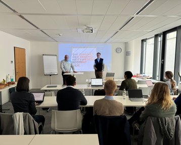 Zwei Personen stehen vorne in einem Konferenzraum und präsentieren. Im Hintergrund ist eine Präsentation mit dem Text „Die Zukunft der Gesundheitswesen beginnt hier“ zu sehen. Teilnehmer sitzen an Tischen mit Laptops.