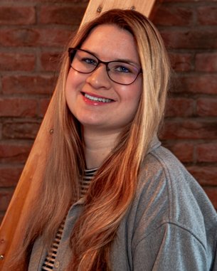 Eine lächelnde Frau mit langen, blonden Haaren und Brille steht vor einer Backsteinwand. Sie trägt ein graues Oberteil und hat eine gestreifte Bluse darunter.