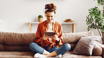 Eine Frau sitzt auf einem Sofa, trägt Kopfhörer und lächelt, während sie ein Tablet in den Händen hält. Sie trägt ein orangefarbenes Oberteil und Jeans. Im Hintergrund sind Pflanzen und ein Regal sichtbar.
