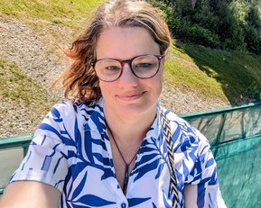 Eine Person mit Brille trägt ein blau-weißes Hemd und lächelt. Im Hintergrund ist eine grüne Landschaft zu sehen.