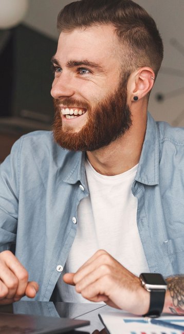 Ein lächelnder Mann mit Bart sitzt an einem Tisch vor einem Laptop. Auf dem Tisch liegen Dokumente und ein Smartphone. Der Raum hat eine moderne Einrichtung.